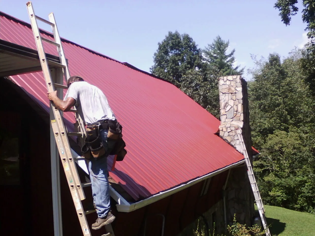 Roofer on a ladder beside a red metal roof and stone chimney — Soffit & Fascia Repair by Roofing Brothers in Blackfoot
